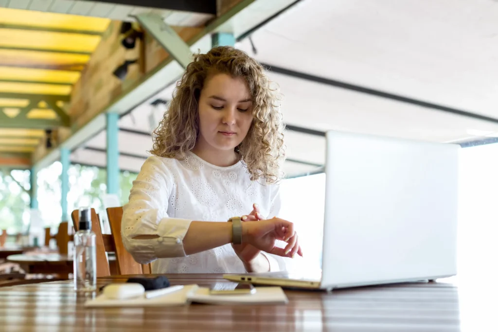 Mujer joven revisando la hora en su reloj inteligente mientras trabaja con su laptop en una terraza.