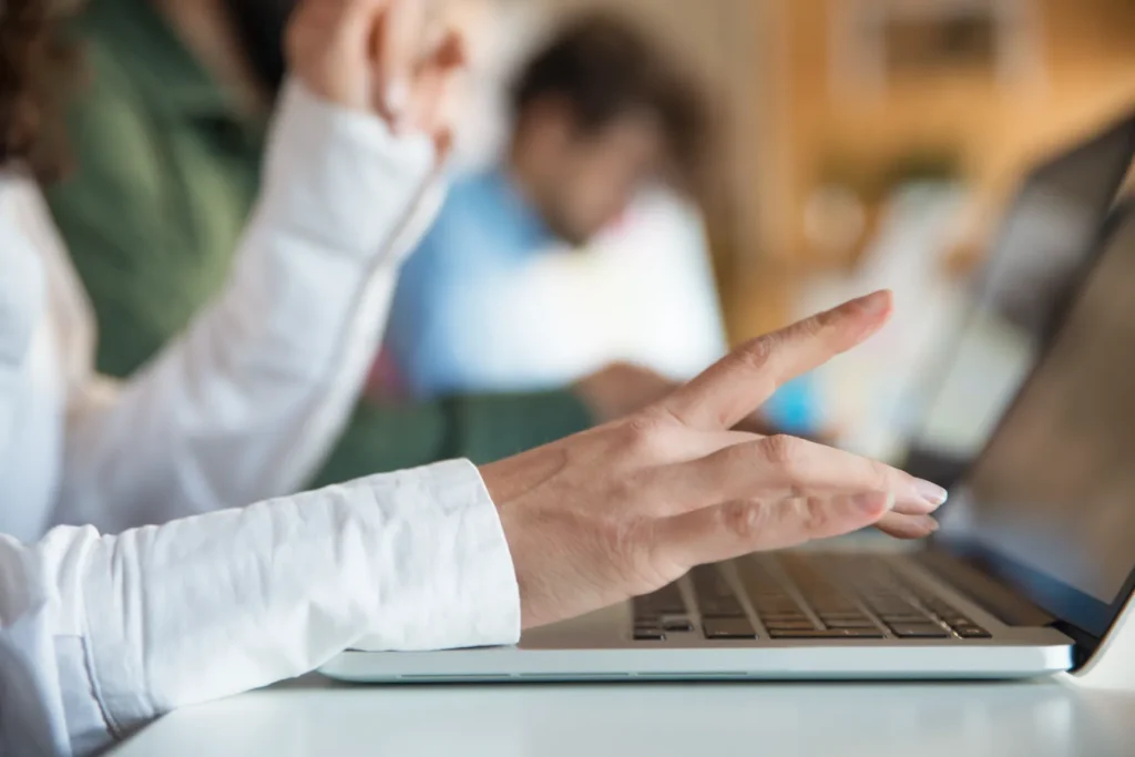 Persona usando una laptop en una mesa de trabajo compartido, con otras personas desenfocadas al fondo.